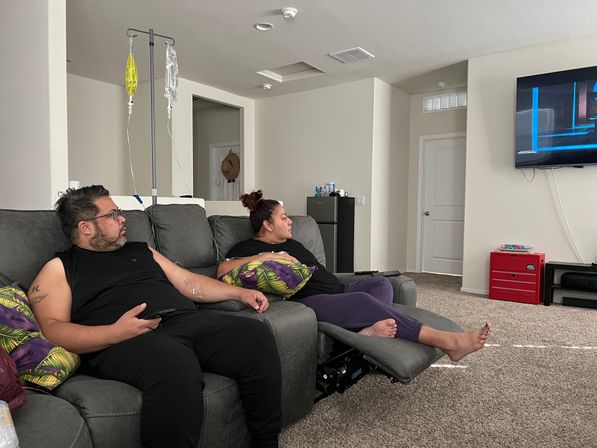 Couple relaxing on a gray sectional couch in a bright home living room, watching wall-mounted TV while one person receives at-home IV infusion from a pole with yellow and clear bags.