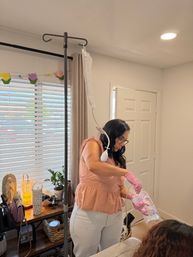 Home caregiver in a peach top wearing pink gloves handling a clear medical fluid bag attached to an IV pole in a bright living room with blinds, potted plant and wooden side table.