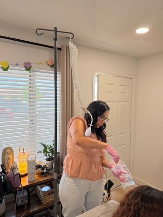 Home caregiver in a peach top wearing pink gloves handling a clear medical fluid bag attached to an IV pole in a bright living room with blinds, potted plant and wooden side table.