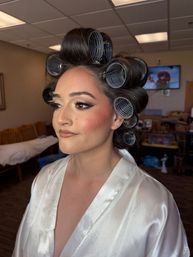 Woman in a white satin robe with large gray hair rollers and glam makeup—winged eyeliner, long lashes and bronzed cheeks—getting ready in a dressing room for bridal beauty prep.
