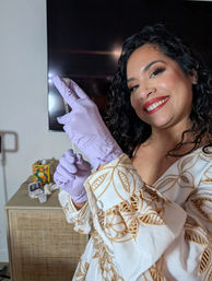 Smiling woman with curly hair in a white embroidered dress wearing lavender gloves and holding a syringe-like vial, posing indoors by a rattan cabinet and wall-mounted TV