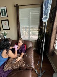 Woman receiving at-home IV infusion on a brown leather couch as a caregiver in pink gloves administers treatment, IV pole and window shutters in a cozy living room