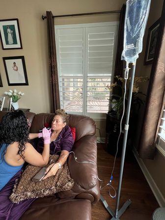 Woman receiving at-home IV infusion on a brown leather couch as a caregiver in pink gloves administers treatment, IV pole and window shutters in a cozy living room