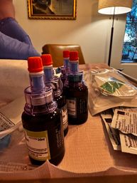 Amber 30ml multi-dose medicine bottles with bright orange caps lined up on a tabletop beside sterile packaging and medication sachets, warm lamp and chair visible in a cozy indoor setting