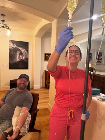 Smiling clinician in pink scrubs and gloves adjusts IV bag while a seated man receives at-home IV infusion in a cozy residential living room.