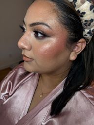 Close-up side profile of a woman showcasing a glam makeup look with bold winged eyeliner, long false lashes, shimmery highlighter and rosy blush, glossy nude lips, leopard-print headband and pink satin robe