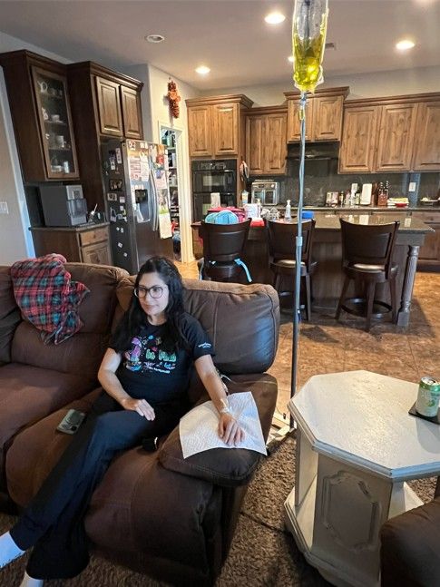 Person relaxing on a brown recliner in a cozy open-plan living room and kitchen, with a yellow IV drip on a stand nearby; wooden cabinets, bar stools, refrigerator with magnets, and a side table with a drink visible.