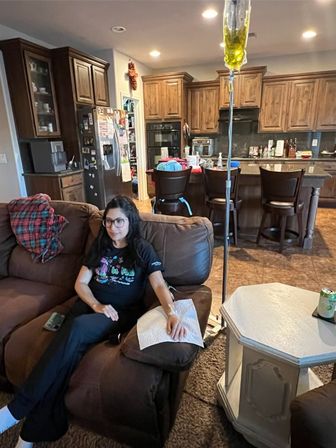 Person relaxing on a brown recliner in a cozy open-plan living room and kitchen, with a yellow IV drip on a stand nearby; wooden cabinets, bar stools, refrigerator with magnets, and a side table with a drink visible.