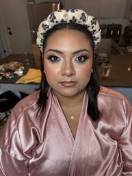 Close-up of a person in full glam makeup—smoky eyes, dramatic lashes, glossy brown lips—wearing a leopard-print padded headband and pink satin robe with a gold necklace, seated in a home kitchen setting.