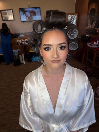 Woman in a white satin robe with large hair rollers and full glam makeup, seated in a dressing room getting ready with salon tools and a TV visible in the background.
