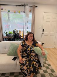 Smiling woman in a floral dress sitting on a beige sofa with green textured pillows receiving an at‑home IV infusion, sunlit window with blinds and tulip garland, entry door and a side table with plants and decor in the cozy living room
