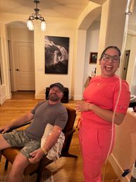 Man seated in a cozy living room chair with an IV line in his arm, smiling; a laughing woman in bright pink scrubs stands beside him, warm wood floors and wall art visible.
