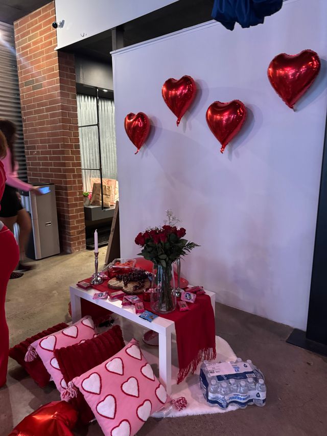 Indoor Valentine's display with red metallic heart balloons on a white backdrop, vase of red roses and snacks on a red-draped table, and pink heart-pattern pillows on the floor beside exposed brick.