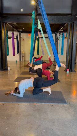 Indoor aerial yoga class with colorful silk hammocks hanging from the ceiling and participants on mats practicing suspended leg and inverted poses