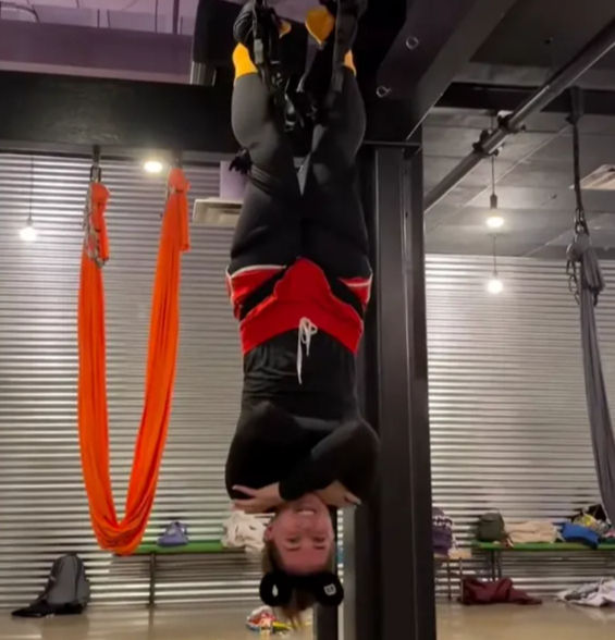 Smiling person hanging upside-down in an indoor aerial fitness studio, feet secured in suspension straps, wearing black leggings and red shorts with bright orange aerial silks and industrial metal wall in the background.