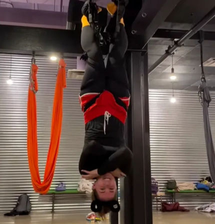 Smiling person hanging upside-down in an indoor aerial fitness studio, feet secured in suspension straps, wearing black leggings and red shorts with bright orange aerial silks and industrial metal wall in the background.