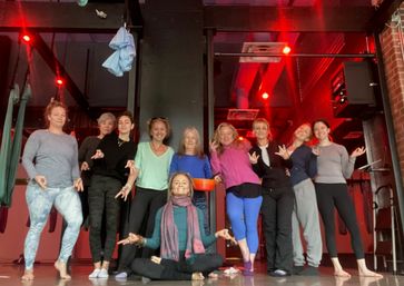 Smiling group of women in an indoor aerial yoga studio posing under red ambient lights with hanging silk hammocks, one woman seated cross‑legged in meditation mudra.