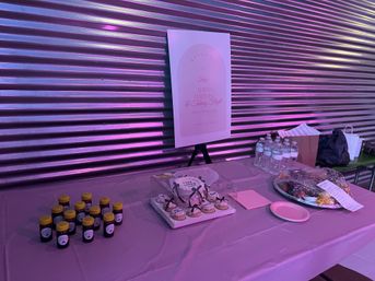 Indoor event table bathed in purple-pink light against a corrugated metal wall, with a welcome poster on an easel, boxed cupcakes with ribbons, rows of small jars with yellow lids, stacked bottled water and wrapped food platters.