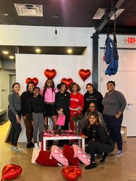 Group of women posing in a fitness studio for a Valentine's Day gathering with red heart balloons, pink heart pillows, a table of roses and treats, and aerial silks hanging from the ceiling.