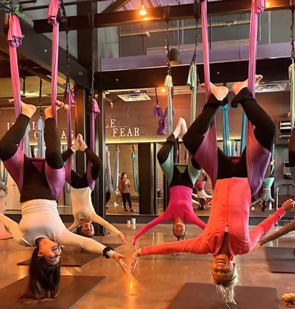Group aerial yoga class in an indoor fitness studio with women suspended upside-down in purple silks, smiling and reaching toward each other.