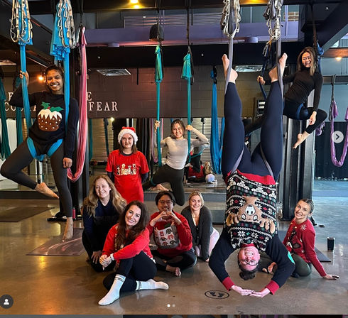 Group in an indoor aerial yoga studio practicing silk hammock poses — several suspended, one upside-down — wearing festive sweaters and a Santa hat.