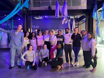 Smiling group of adults in an aerial yoga class inside an urban fitness studio with colorful silk hammocks hanging from exposed beams and purple‑blue mood lighting