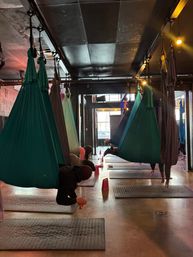 Indoor aerial yoga studio with rows of teal and gray fabric hammocks suspended from the ceiling, yoga mats on the floor and participants practicing suspended poses in a dimly lit room.