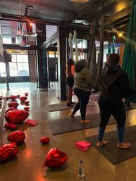 Urban aerial yoga studio interior with suspended silks and mats, people preparing for class and red heart balloons scattered on the polished concrete floor near large industrial windows.