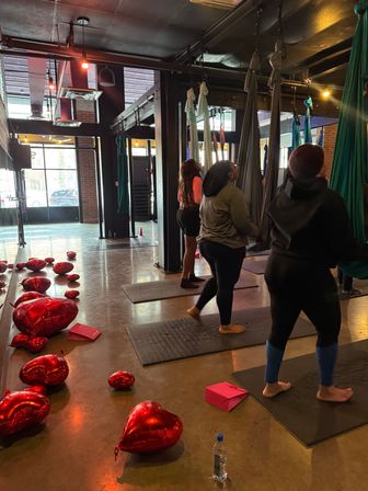 Urban aerial yoga studio interior with suspended silks and mats, people preparing for class and red heart balloons scattered on the polished concrete floor near large industrial windows.