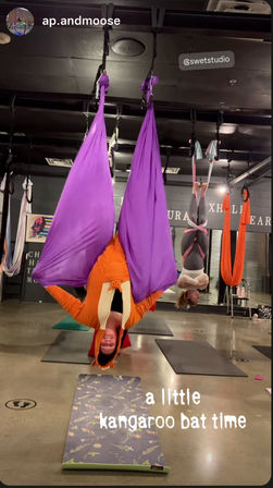 Smiling person in an orange kangaroo onesie hanging upside down in a purple aerial yoga hammock during an indoor aerial-silks class with yoga mats.