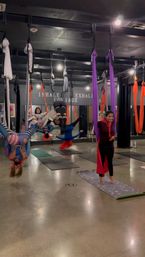 Group aerial yoga class in an indoor fitness studio with colorful silk hammocks, students practicing inversions and standing poses on mats.