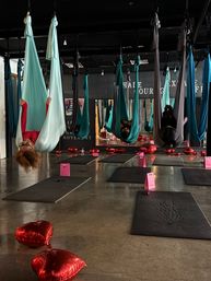 Indoor aerial yoga class with teal and gray silk hammocks hanging from the ceiling, participants suspended upside down over black mats, pink gift bags and red heart-shaped balloons scattered across a polished concrete floor and mirrored wall in the background.