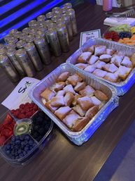 Catering table with trays of powdered beignets, mixed berry and fruit platters, and rows of slim beverage cans on a dark wood table under blue event lighting.