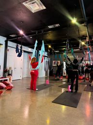 Group aerial yoga class in an indoor fitness studio with turquoise silk hammocks suspended from the ceiling, participants on mats reaching upward in a lively group workout.