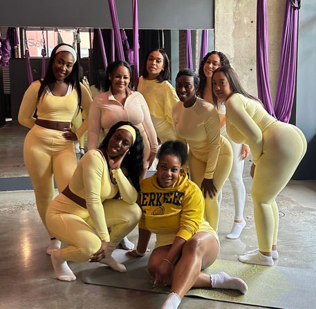 Eight women in coordinating yellow activewear smiling and posing on a mat inside an aerial yoga fitness studio with purple silks, a mirrored wall and concrete floor