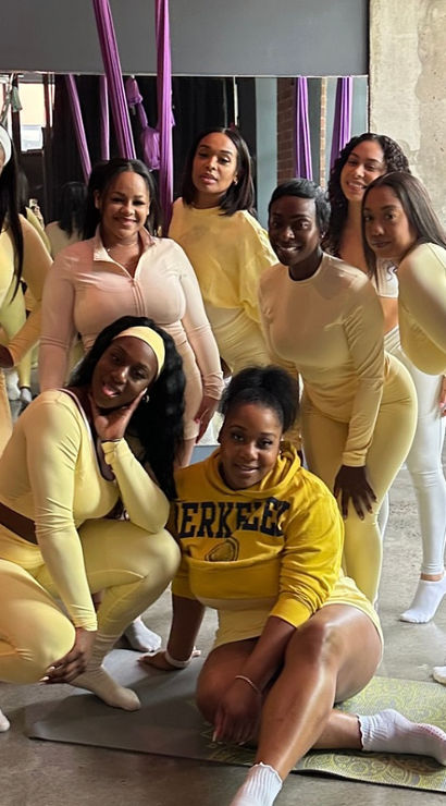 Eight women in coordinating yellow activewear smiling and posing on a mat inside an aerial yoga fitness studio with purple silks, a mirrored wall and concrete floor