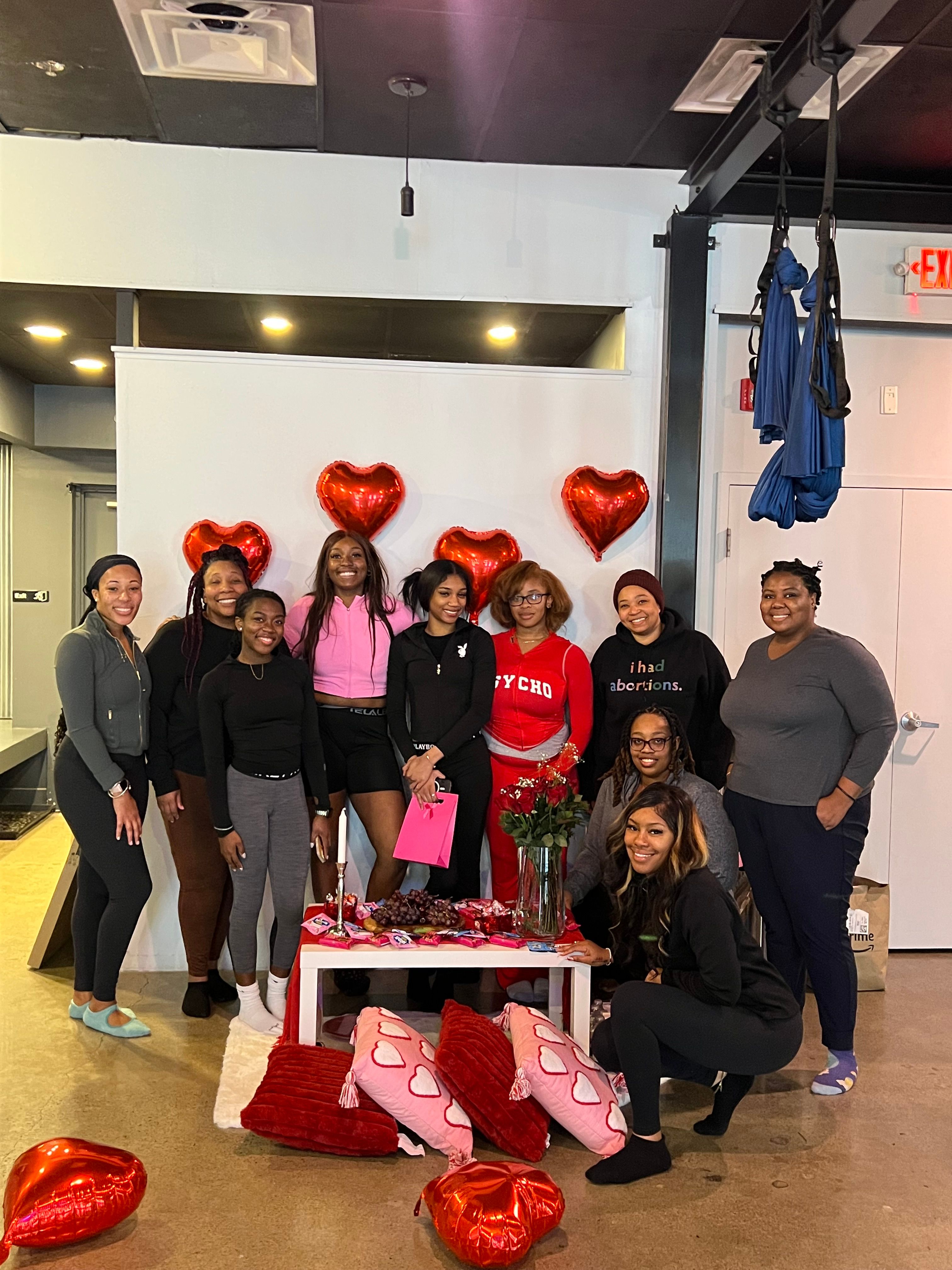 Group of women smiling in an indoor studio at a Valentine’s-themed gathering, posing in front of red heart balloons with a table of roses, snacks, a pink gift bag and pink heart pillows on the floor.