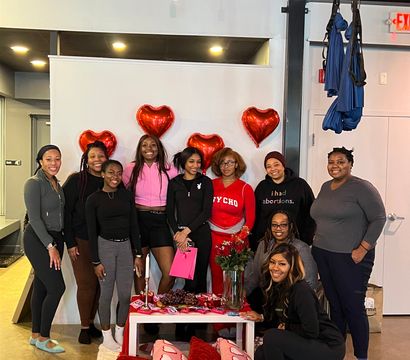 Group of women smiling in an indoor studio at a Valentine’s-themed gathering, posing in front of red heart balloons with a table of roses, snacks, a pink gift bag and pink heart pillows on the floor.