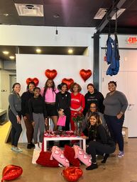Smiling group of women in an indoor fitness studio posing with red heart balloons, Valentine-themed pillows and treats on a table and blue aerial silks overhead.