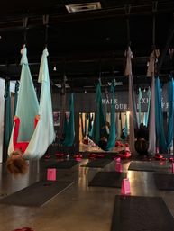 Aerial yoga studio interior with teal and gray silk hammocks hanging from the ceiling, participants suspended upside-down over black mats, pink gift bags and red balloons on the polished concrete floor.