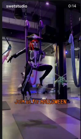 Spooky Halloween aerial yoga scene: person in a skeleton bodysuit and pirate hat suspended on purple aerial silks inside a dim, purple-lit studio with hanging silks, floor mats, and festive decorations.