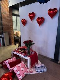 Valentine's Day indoor display: red heart foil balloons on a white backdrop, vase of red roses and snacks on a small table, pink heart-pattern pillows on the concrete floor — romantic event decor.