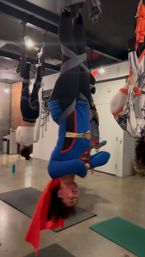 Group doing aerial yoga inversions in a fitness studio, one participant suspended upside down in a blue superhero-style outfit with a red cape.
