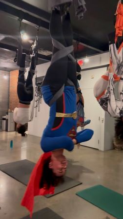 Group doing aerial yoga inversions in a fitness studio, one participant suspended upside down in a blue superhero-style outfit with a red cape.
