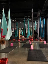 Aerial yoga studio with teal and gray silk hammocks suspended from the ceiling, students inverted in the hammocks above black mats and pink gift bags, red heart balloons on the polished concrete floor.