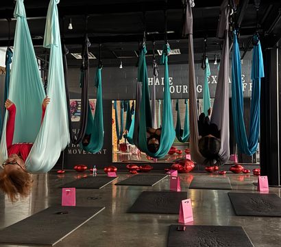 Aerial yoga studio with teal and gray silk hammocks suspended from the ceiling, students inverted in the hammocks above black mats and pink gift bags, red heart balloons on the polished concrete floor.