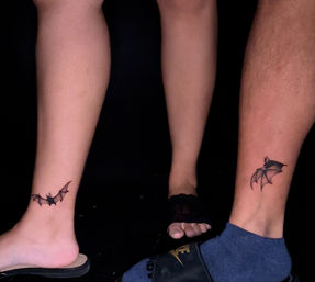 Close-up of two lower legs and feet against a black background showing matching minimalist black bat ankle tattoos—tiny flying bat designs on each ankle.