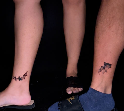 Close-up of two lower legs and feet against a black background showing matching minimalist black bat ankle tattoos—tiny flying bat designs on each ankle.