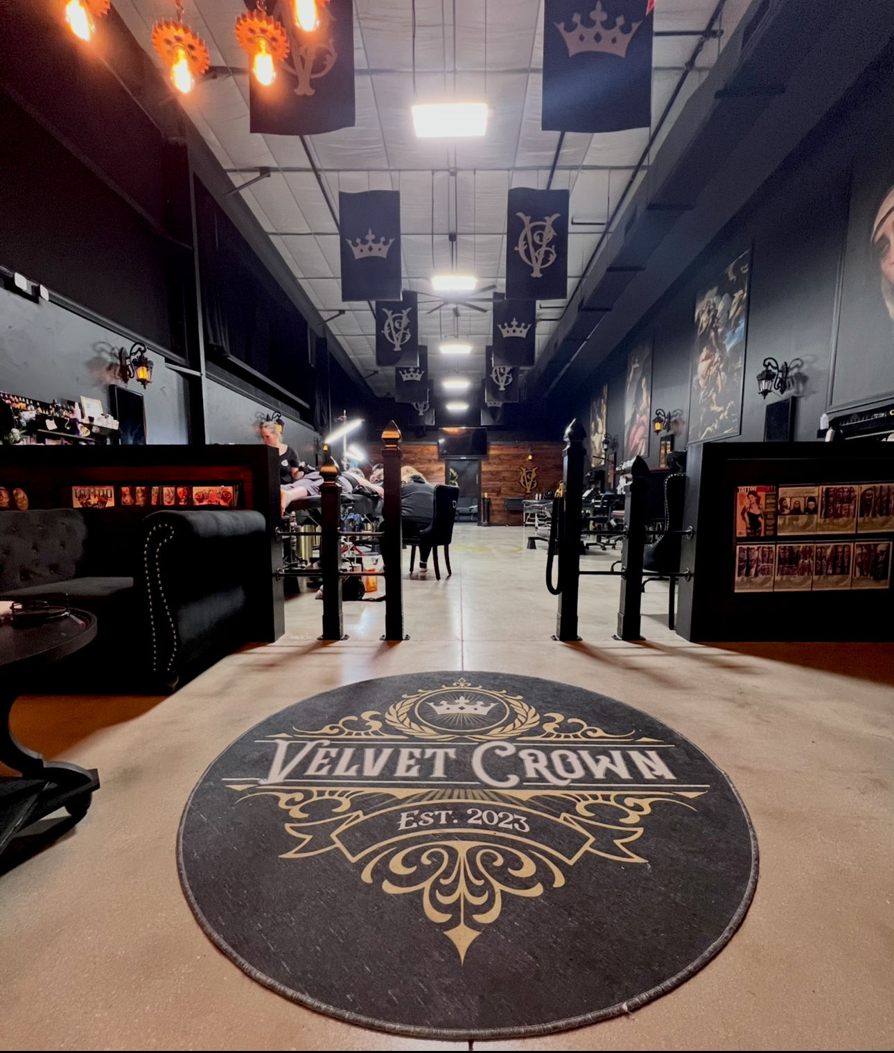 Wide-angle tattoo shop interior with industrial ceiling and hanging black banners, round ornate entrance rug showing Est. 2023, tattoo stations with artists at work, dark lounge seating and large framed artwork along the walls.