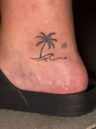 Ankle tattoo of a minimalist palm tree and wave with a small sun and tiny heart, shown on skin above a black slide sandal